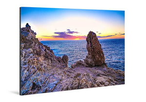 Tableau mural représentant un coucher de soleil sur la mer entre falaises rocheuses, avec un grand rocher dressé face à l’horizon