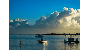 Peaceful seascape at sunset with dramatic clouds, small boats, and a child standing in shallow water