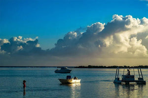 Fine art photo of boats on calm water at sunset with dramatic clouds and a person standing in shallow water