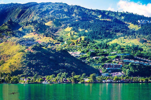 Lush green mountain landscape with scattered houses reflected in calm lake under blue sky