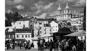 Black and white photo of a lively village square with a statue, historic buildings, and people gathering.