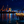 Night harbor scene with illuminated dock restaurant, city lights, cranes, and starry sky