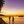 people on tropical beach at sunset with palm trees, boats, and wooden pier reflected on calm ocean