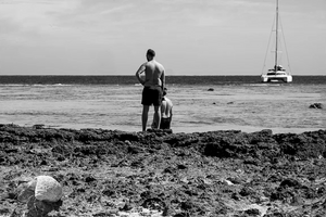 Black and white photo of two men on a rocky beach, yacht in the ocean, large seashell in foreground.